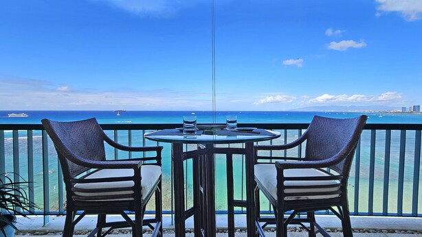 A balcony with two wicker chairs and a table, overlooking a clear blue sea with boats and a distant cityscape. - RELAX w/ Direct Beach Access High Flr. Sans Souci 2BD/2BA🌴Stunning Ocean Views  (Honolulu)