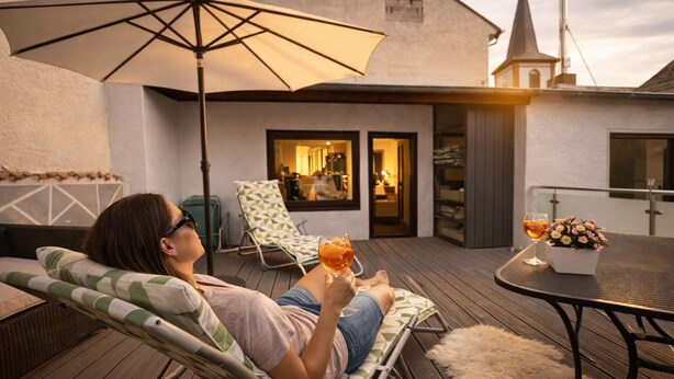 A person relaxing on a lounge chair with a drink, a table with a flower vase, and a view of a building with a tower. - 170 qm2 Großes, Gemütliches Haus mit Kamin und Klimaanlage. Hunde Erwünscht! (Kestert)