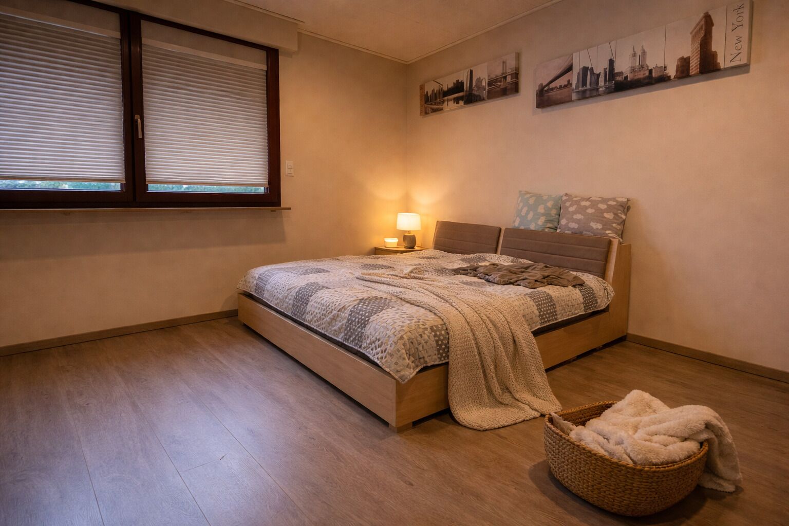 A bedroom with a bed, a window with blinds, a lamp, and a wall with cityscape artwork.