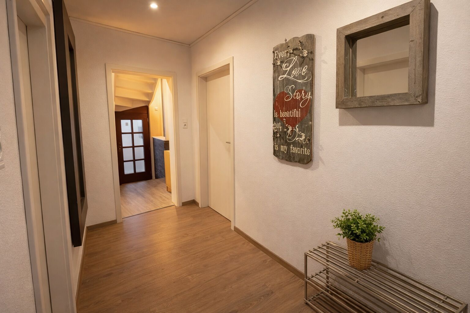 Un pasillo con piso de madera, una puerta, un espejo fijado a la pared y un letrero decorativo.
