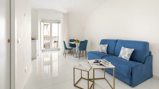 Una sala de estar moderna con un sofá azul, una mesita de centro pequeña y una zona de comedor con una mesa blanca y sillas azules.