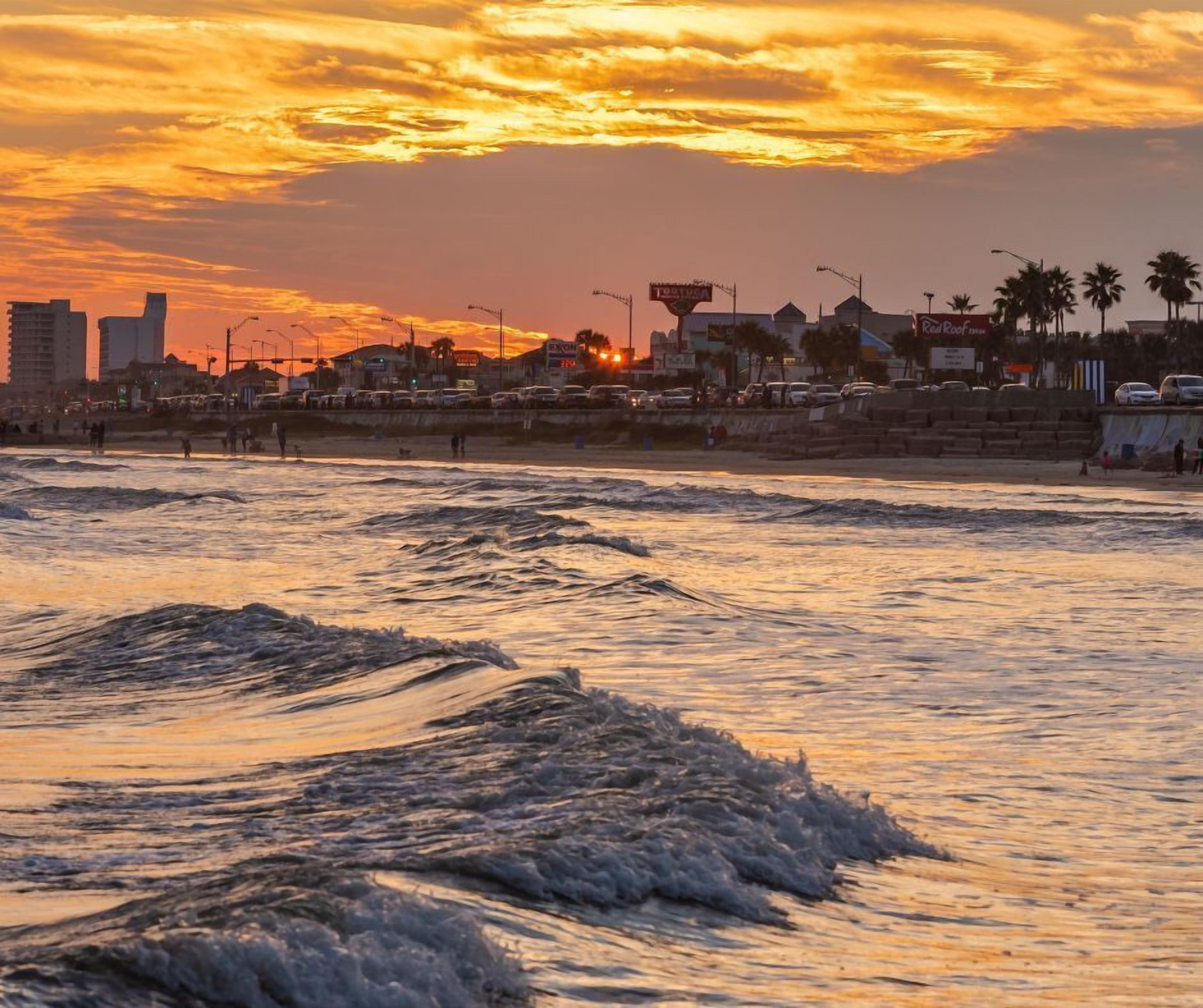A coastal cityscape at sunset with waves crashing onto the shore, buildings, and palm trees in the background.