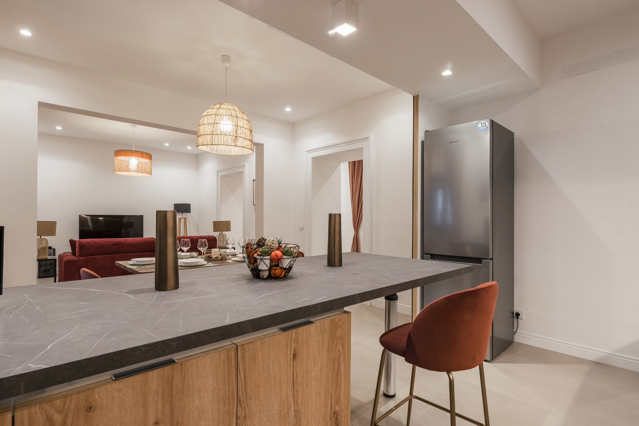 A modern kitchen with a marble countertop, a stainless steel refrigerator, and a dining area with a red sofa and a table.