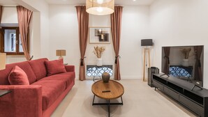 En moderne stue med en rød sofa, et rundt træbord, et fjernsyn og en pejs med dekorative genstande.