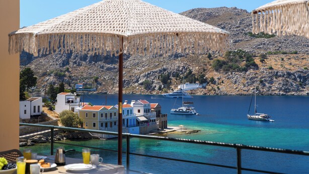 A balcony with a table set for breakfast overlooking a coastal town with buildings and boats. - Villa in Symi, with sea view (simi)