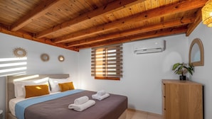 A bedroom with a wooden ceiling, a bed with a brown quilt, white pillows, and a blue blanket. There is a wooden cabinet with a plant on top, a window with blinds, and an air conditioning unit on the wall. - Villa in Symi, with sea view (simi)