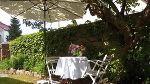 Un patio al aire libre con una mesa y sillas blancas, un paraguas color beige y un florero con flores.