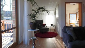 Un salon avec une grande fenêtre, une plante en pot, un canapé gris, un tapis rouge et une petite table.