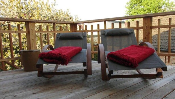 Deux fauteuils de jardin, recouverts de plaids rouges, sont placés sur une terrasse en bois, avec une clôture en bois en arrière-plan.