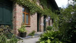 En byggnad av tegel med gröna fönsterluckor, en liten veranda och en trädgård med olika växter och blommor.