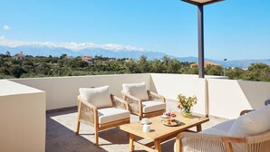 A terrace with wicker furniture, a table with a vase of flowers, and a view of mountains in the distance.