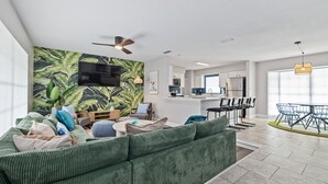 A modern living room with a green sofa, a flat-screen TV, a ceiling fan, and a kitchen in the background. - Pickleball Paradise - Heated Pool-Hot Tub-Game Room (Tampa)
