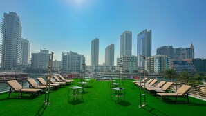 Une terrasse sur le toit, agrémentée d’une pelouse artificielle verdoyante, de chaises longues et de tables, offrant une vue sur un paysage urbain dominé par des immeubles de grande hauteur.