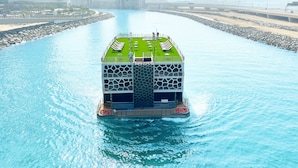 Un bateau doté d’un pont vert et d’un atrium central, entouré d’eau, avec un paysage urbain en arrière-plan.