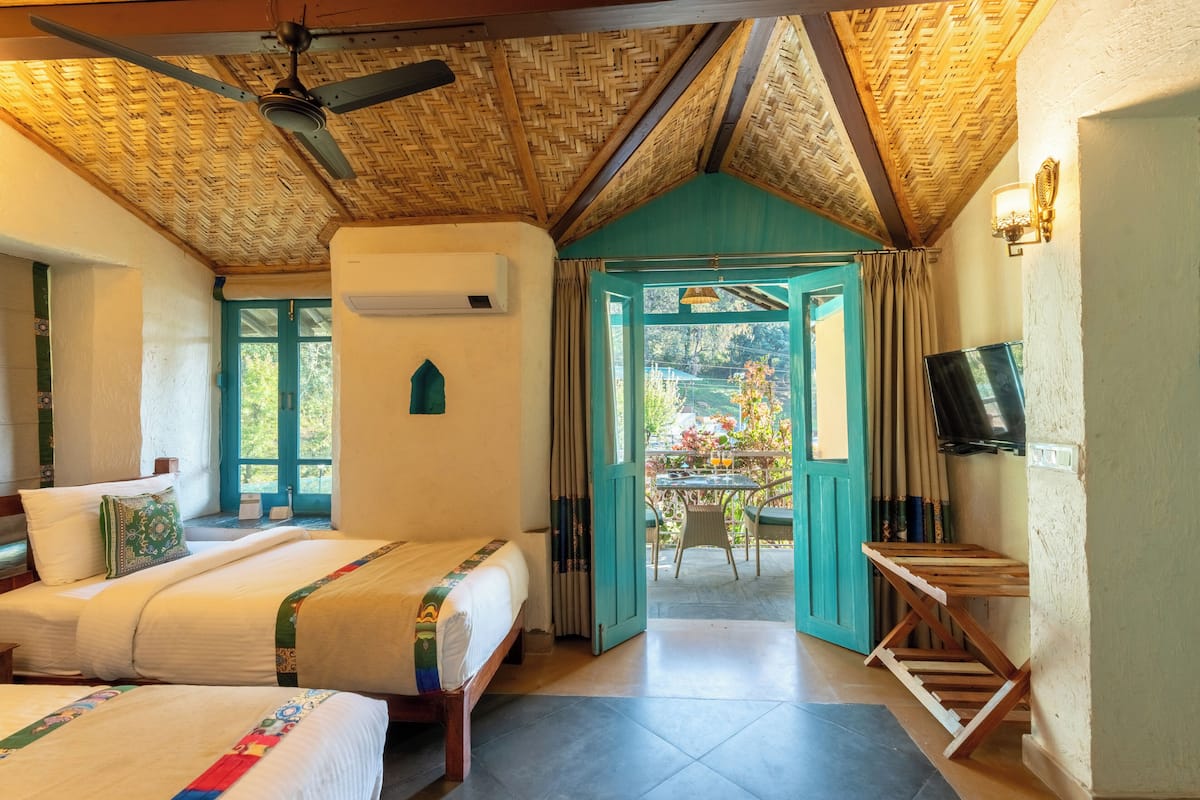 A room with two beds, a ceiling fan, a TV, and a view of a garden through a door.