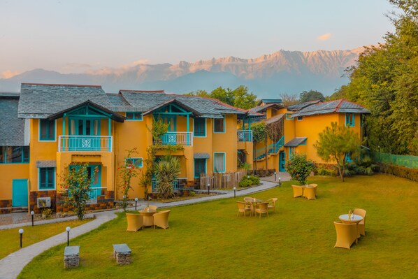 A row of yellow buildings with blue accents, a grassy courtyard with outdoor seating, and a mountain backdrop. - Tree of Life Birdsong Chalets (Dharamshala)