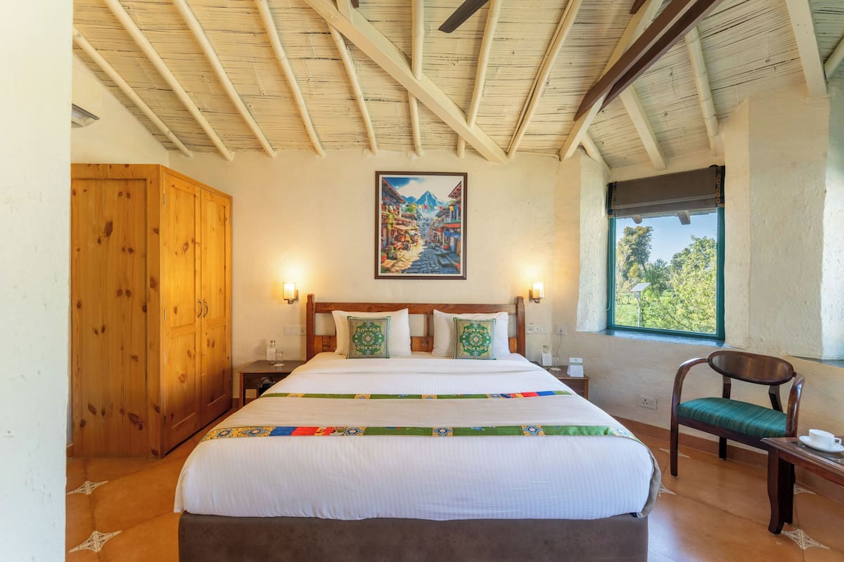 A bedroom with a wooden ceiling, a bed with a colorful quilt, a wooden wardrobe, a chair, and a window with a view of trees.