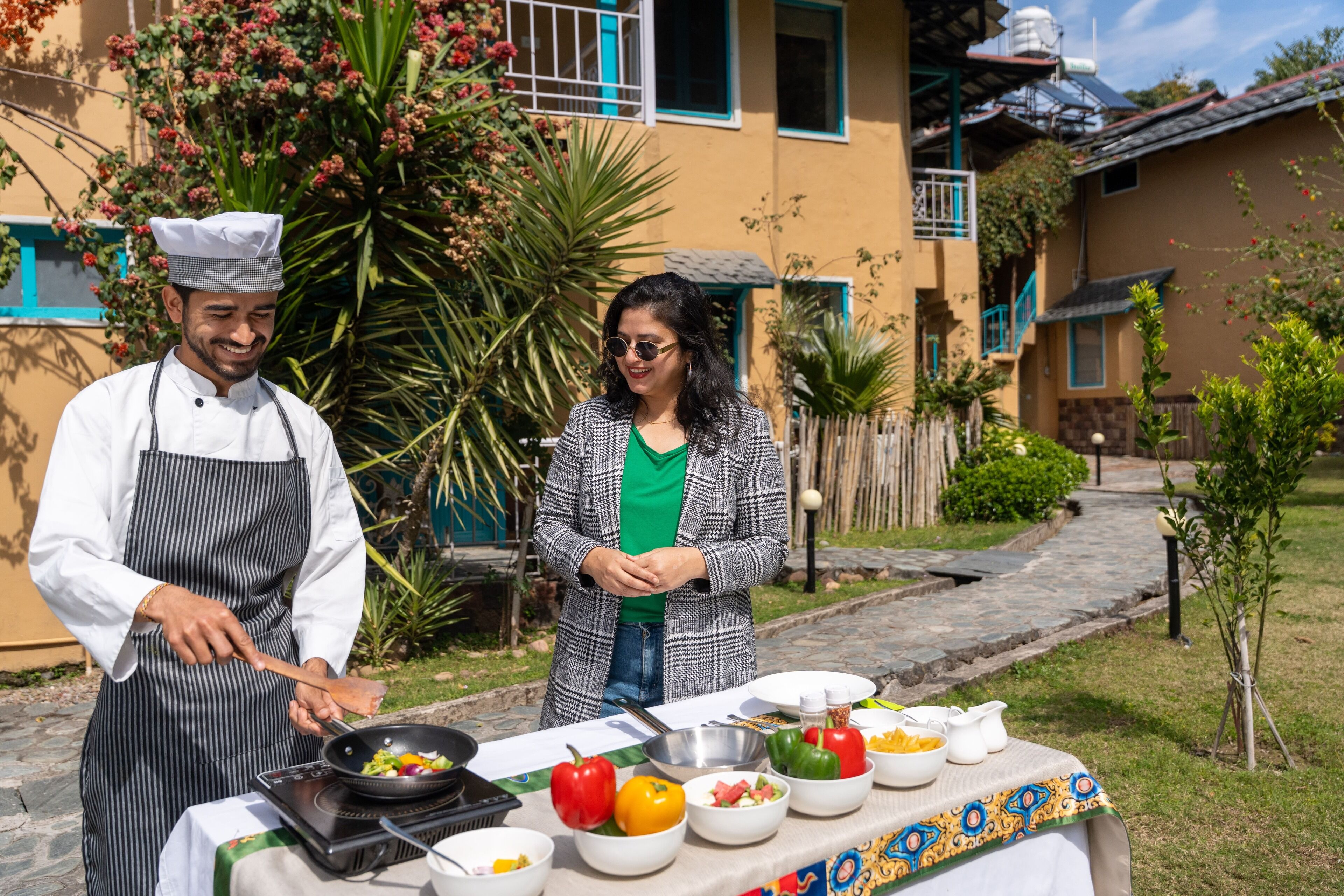 Due persone cucinano all'aperto, con un tavolo pieno di verdure e utensili da cucina.