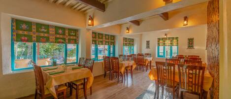 A dining area with multiple tables and chairs, large windows with decorative panels, and wooden beams on the ceiling.