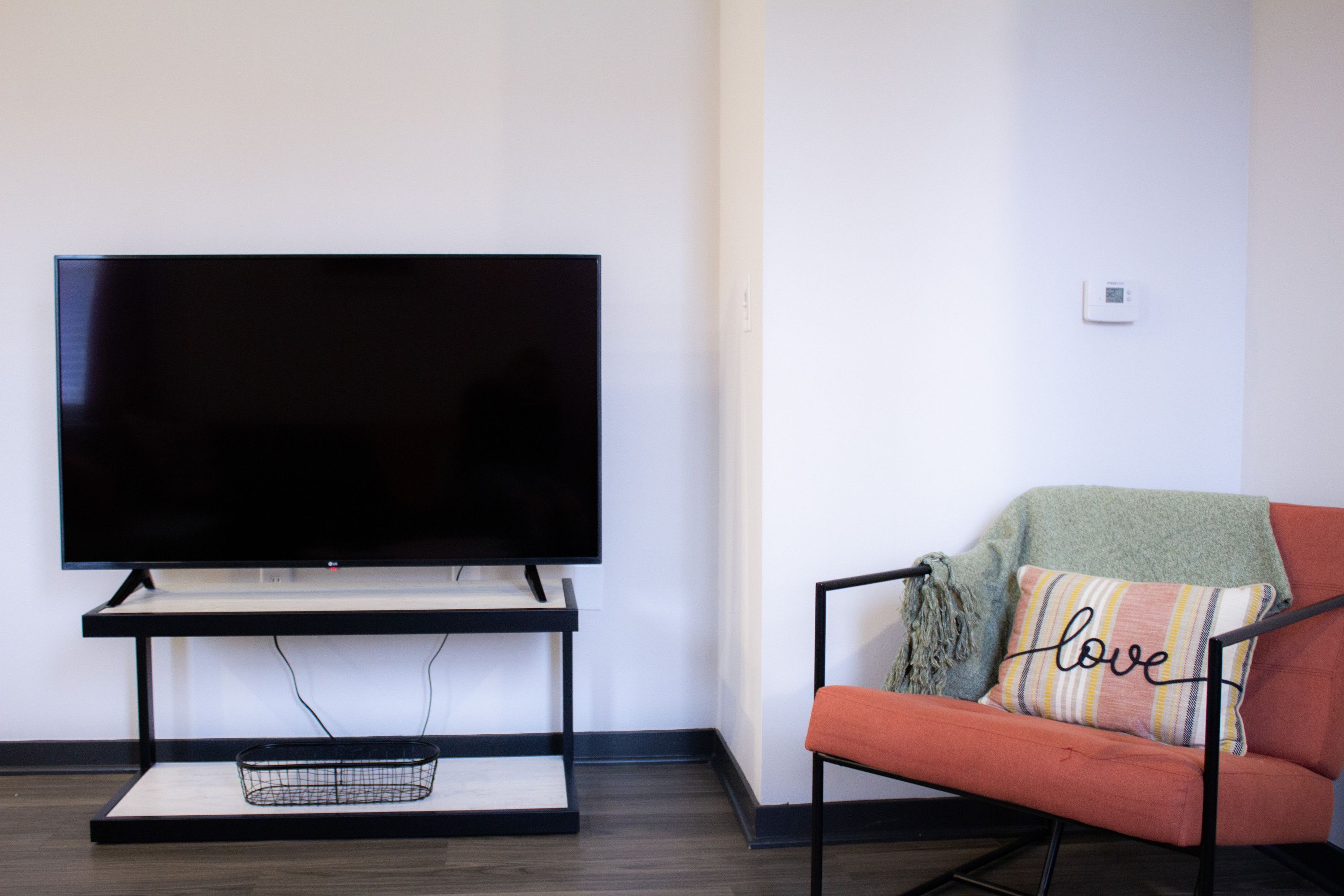 A modern living room with a flat-screen TV on a stand, a red sofa with a 'love' pillow, and a small black basket on the floor.