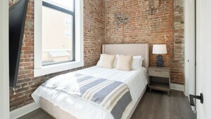 Quarto com parede de tijolos, uma janela grande, cama com manta listrada, mesa de cabeceira com abajur e uma porta.