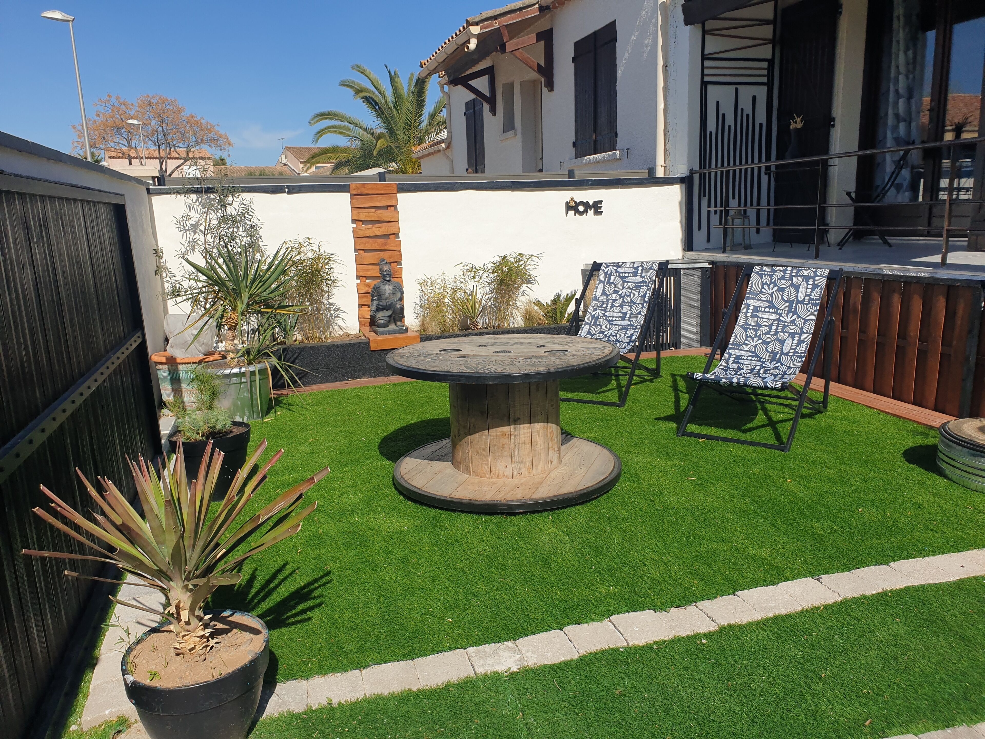 A patio area with a round table and two chairs, surrounded by artificial grass and potted plants.