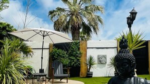 A statue of a seated figure in a garden setting with a palm tree, outdoor furniture, and a building in the background. - Villa Nyvioni, Proche de la mer Avec Piscine et SPA (Agde)
