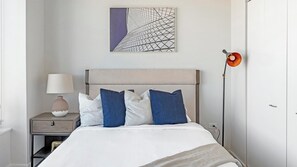 Une chambre à coucher bien rangée, avec un lit, une table de chevet dotée d’une lampe, une œuvre d’art abstraite accrochée au mur et un lampadaire.
