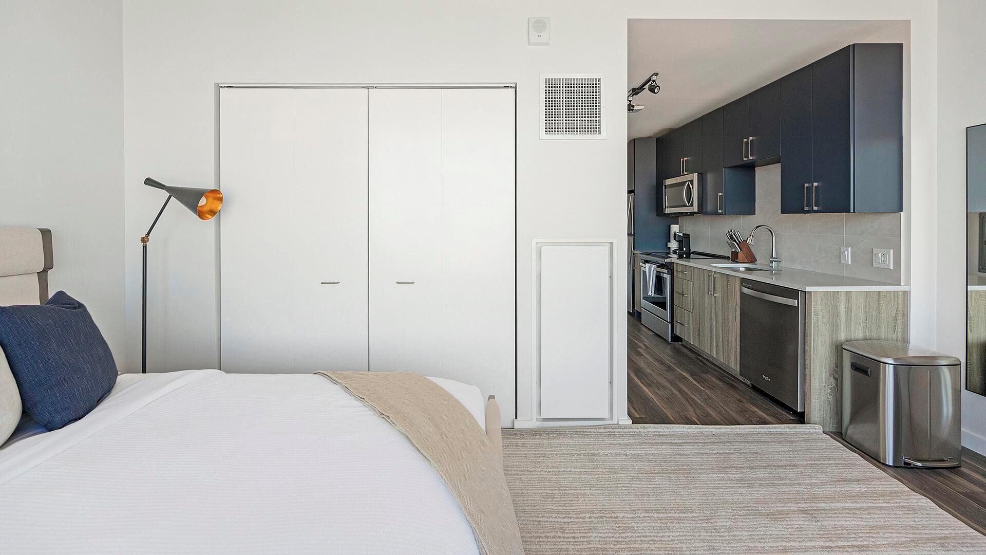 Une chambre avec un lit, une lampe de chevet et une armoire. À côté se trouve une petite cuisine équipée d’un four à micro-ondes, d’une cuisinière et d’un évier.