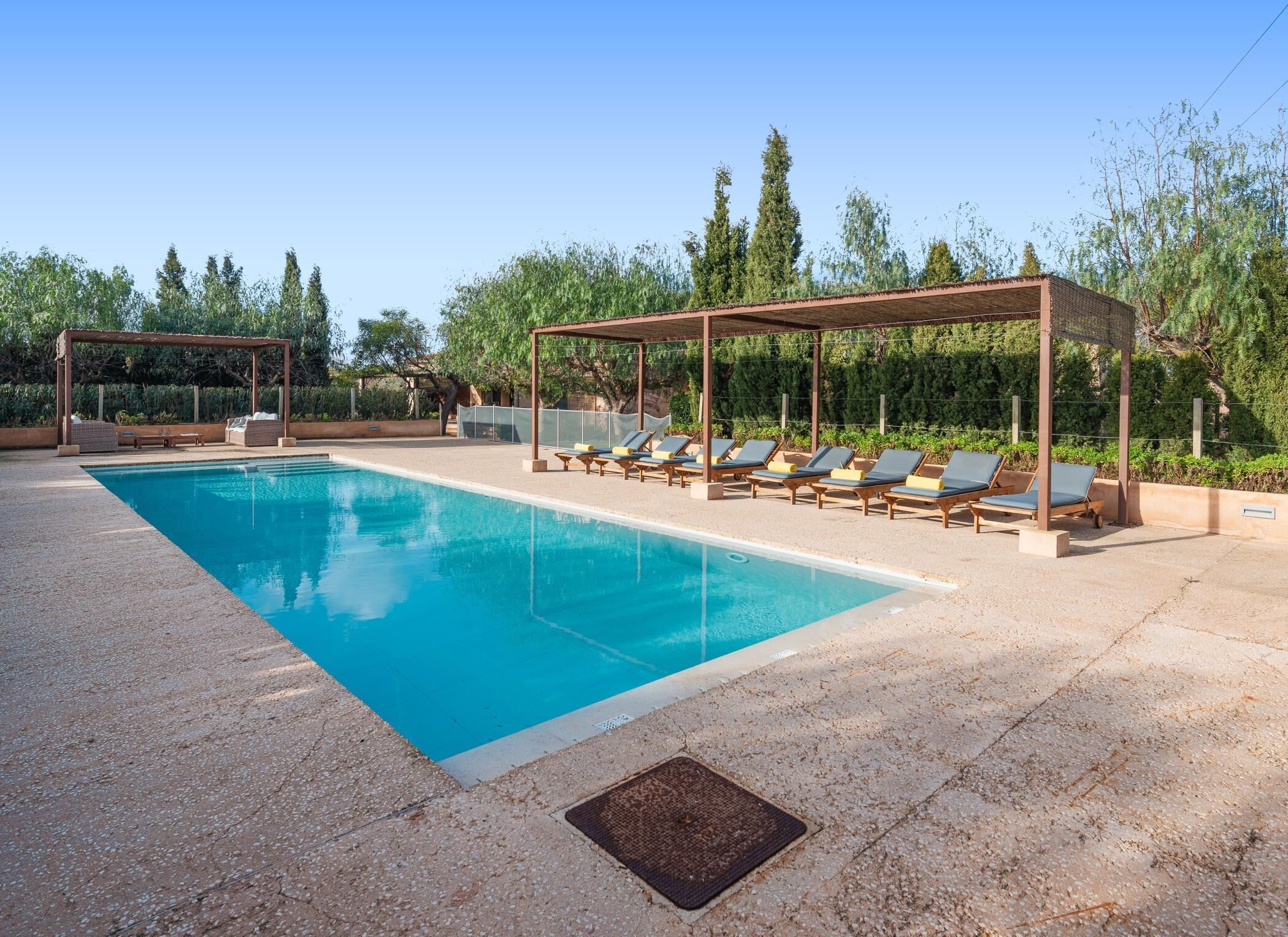 A rectangular swimming pool with clear blue water, surrounded by a pebble deck. There are lounge chairs under pergolas, and the area is bordered by trees and shrubs.