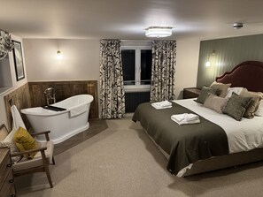 A bedroom with a large bed, a sitting area with a chair, a bathtub, and a window with curtains. - White Lion Hotel (Worcester)