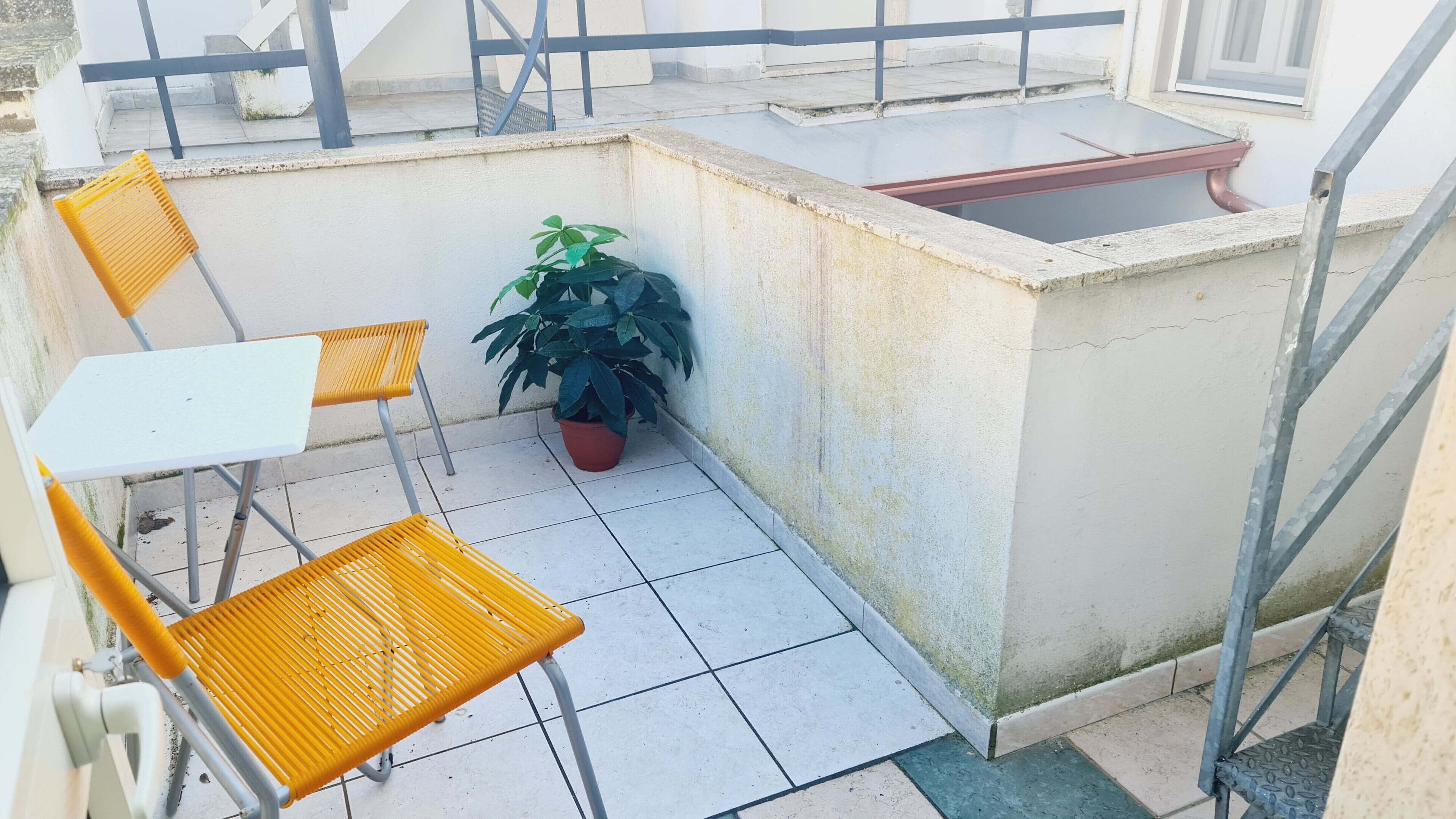 A small outdoor patio with a white table and two yellow chairs, a potted plant, and a concrete sink.
