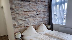 Een slaapkamer met een stenen muur, twee bedden, een nachtkastje met een lamp en een raam met jaloezieën.