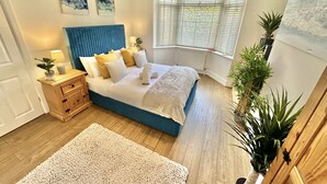 Un dormitorio con cabecera azul, ropa de cama blanca, mesita de noche de madera, alfombra blanca y un ventanal con persianas.