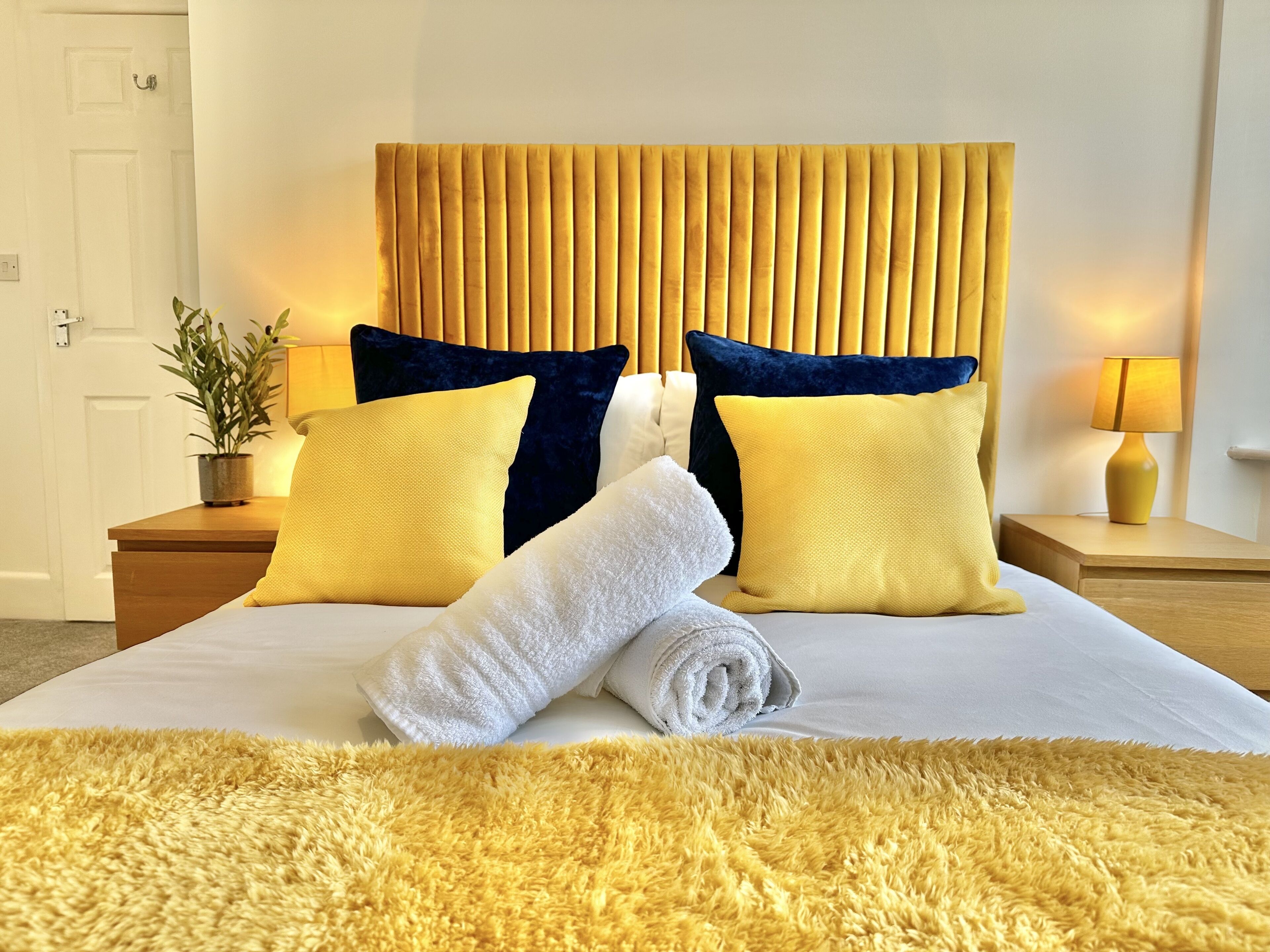 Een net opgemaakt bed met een opgerolde handdoek, een zachtgeel vloerkleed en een hoofdbord met verticale latten.