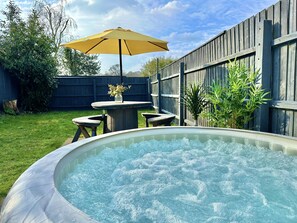 An outdoor hot tub area with a wooden fence, a table with an umbrella, and a grassy lawn.