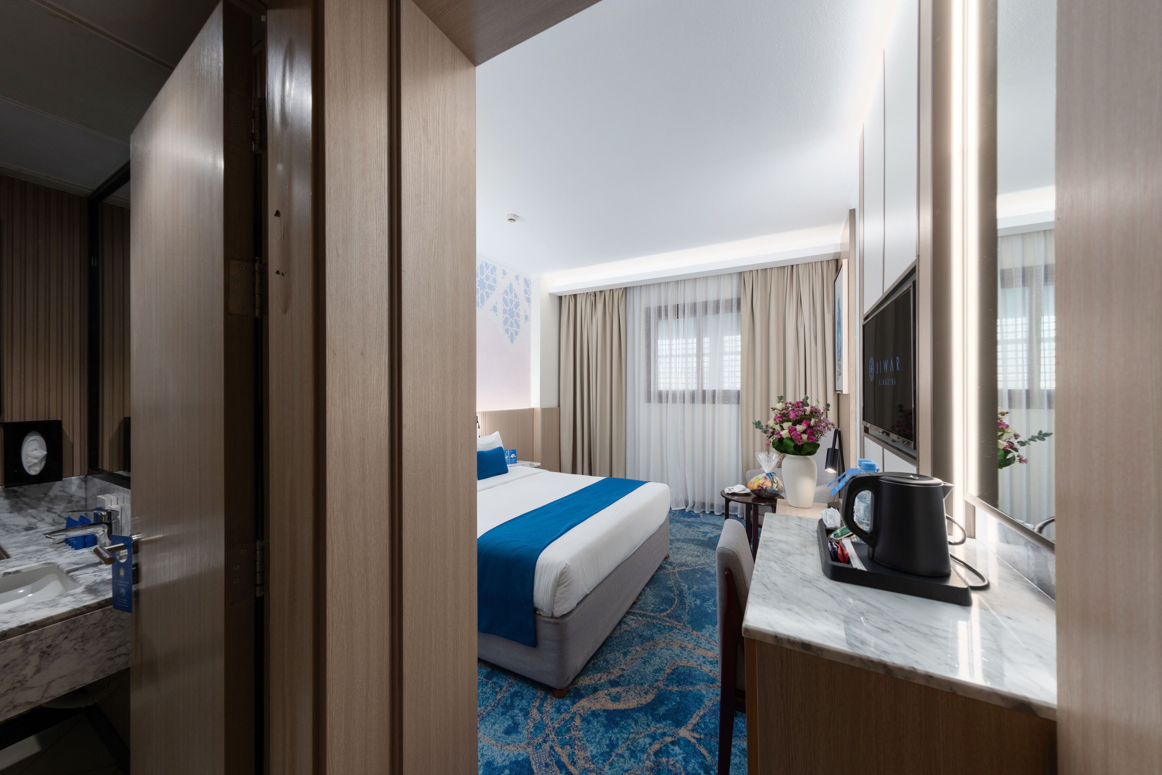 A hotel room with a bed, a desk with a coffee maker, a TV, and a bathroom with a sink and mirror.