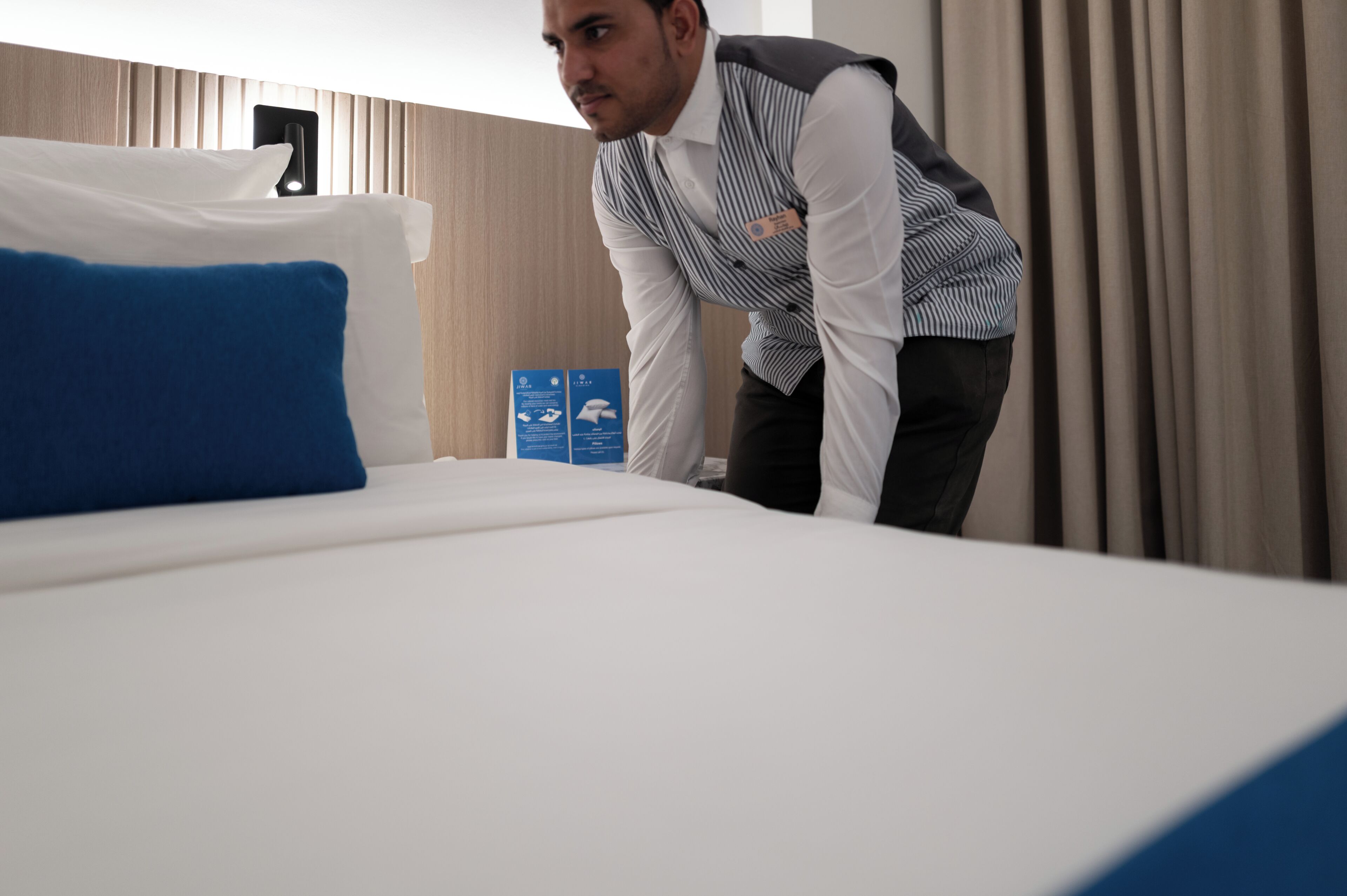 Une chambre d’hôtel avec un lit, des oreillers, un coussin décoratif bleu, une tête de lit en bois et un panneau d’information bleu.