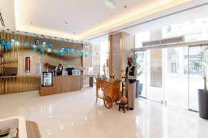 A hotel reception area with a wooden reception desk, a cart with a vase, and a large window with a view of the street.
