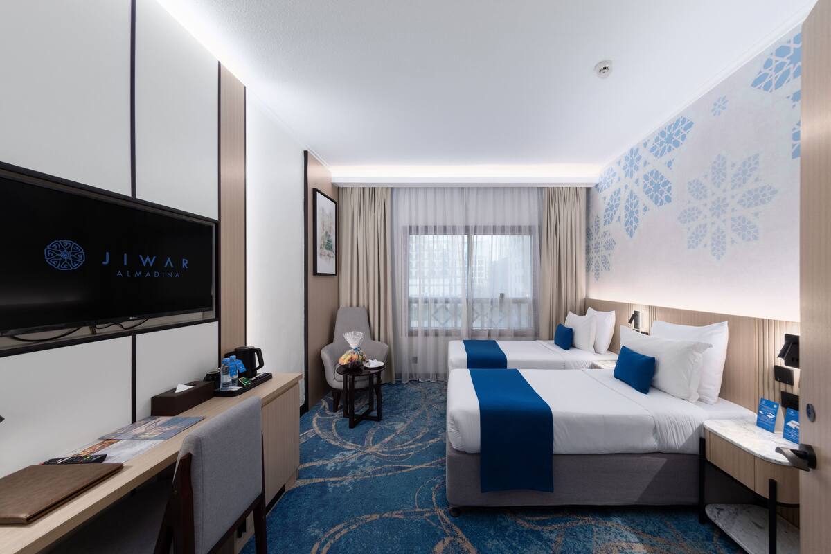 Hotelkamer met flatscreen televisie, twee bedden met blauwe accenten, een bureau met stoel en een kleine tafel met lamp.