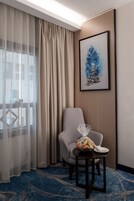 Habitación de hotel con ventana, una silla, una mesita con frutero y una obra de arte enmarcada en la pared.