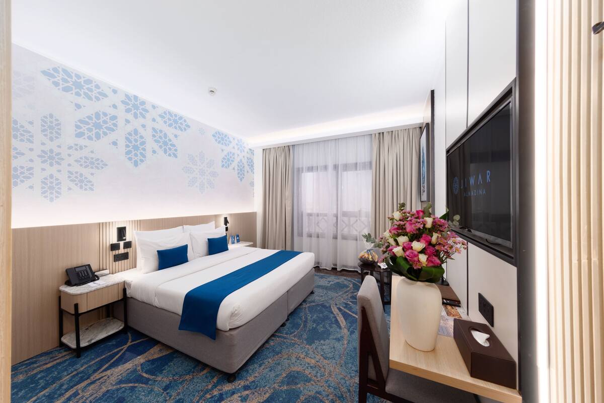Een hotelkamer met een bed, nachtkastje, televisie, bureau, stoel en een decoratieve wand.