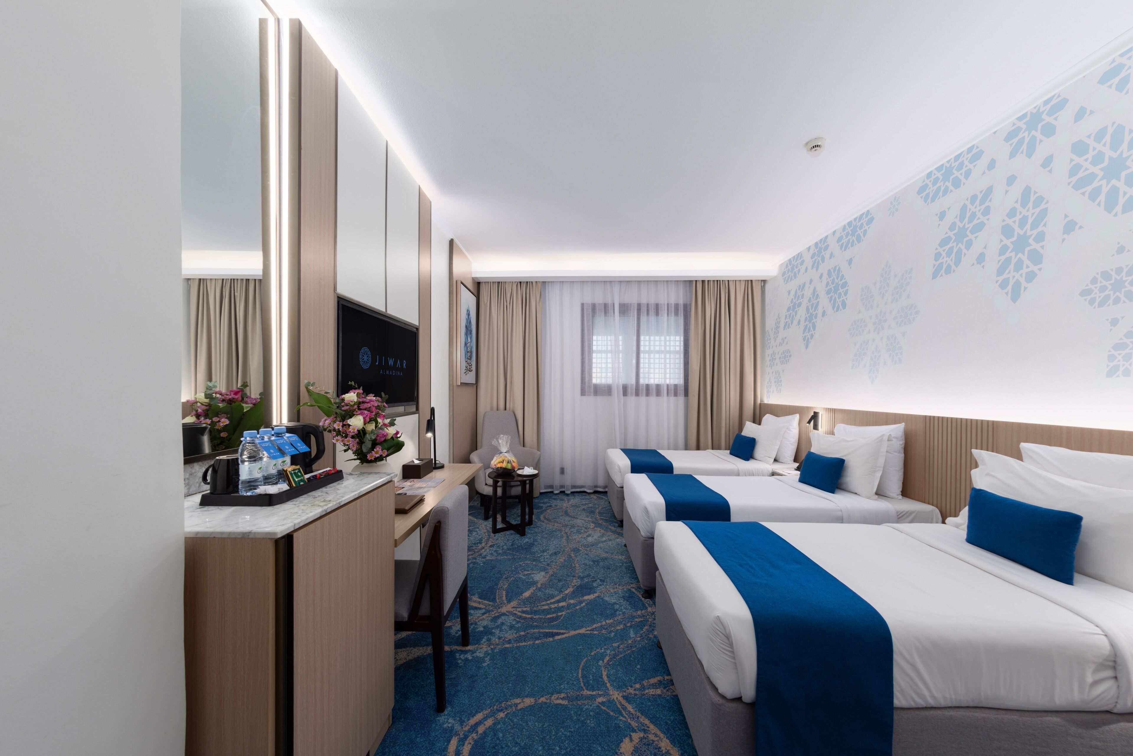 A hotel room with two beds, a desk, a chair, a TV, and a window with curtains.