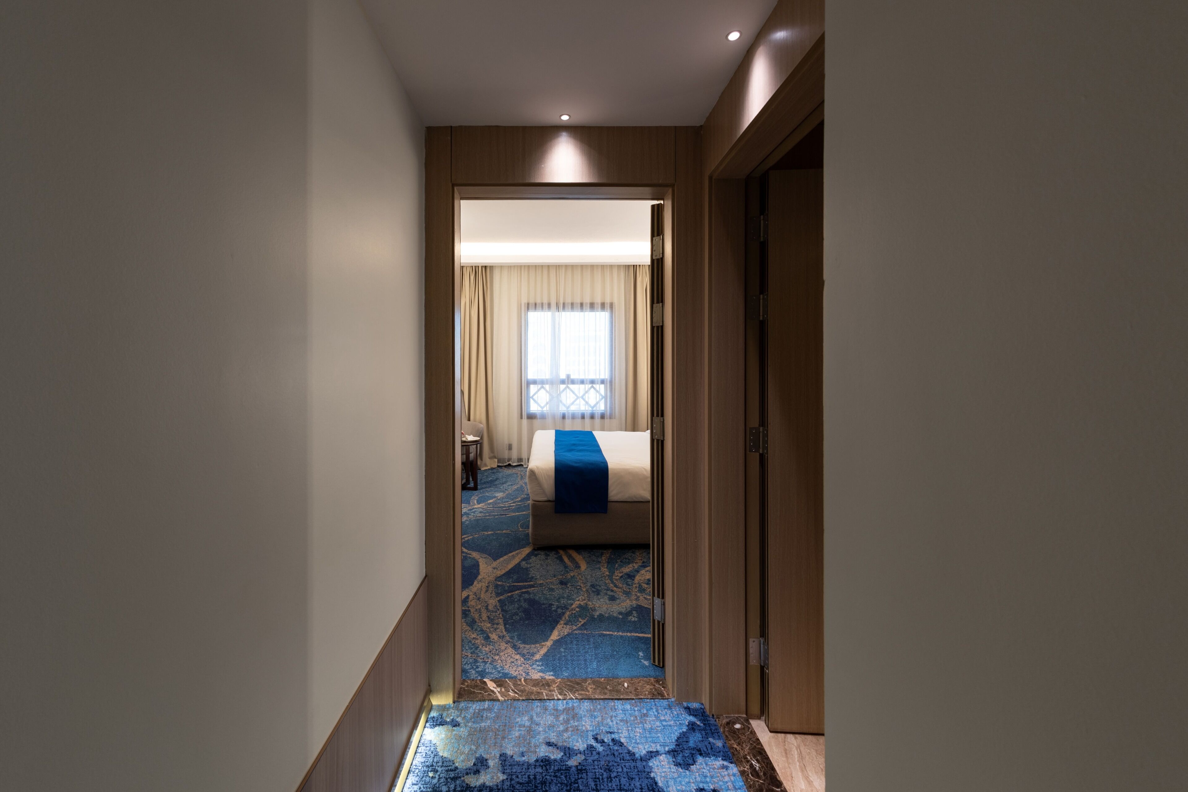 A hotel hallway leading to a room with a bed, a window with curtains, and a blue patterned carpet.