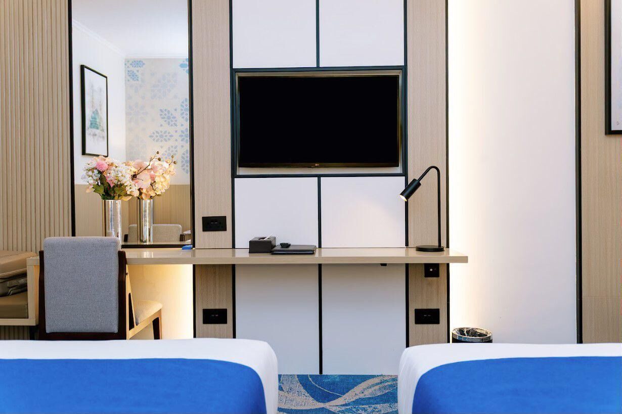 Une chambre d’hôtel comprenant un lit, un bureau avec un téléviseur à écran plat, un vase avec des fleurs et une lampe.