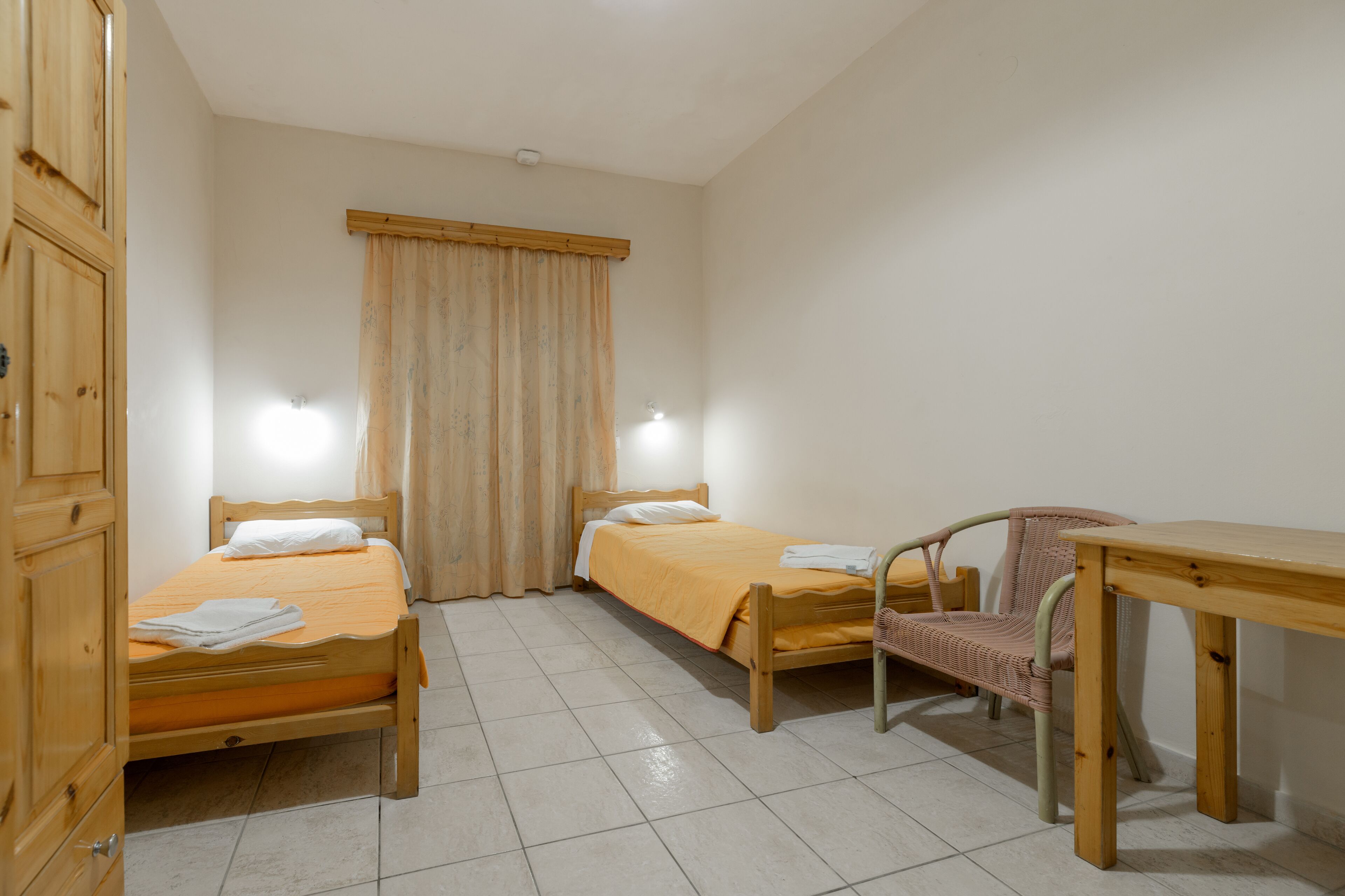 A small, clean room with two wooden beds, a chair, and a table.