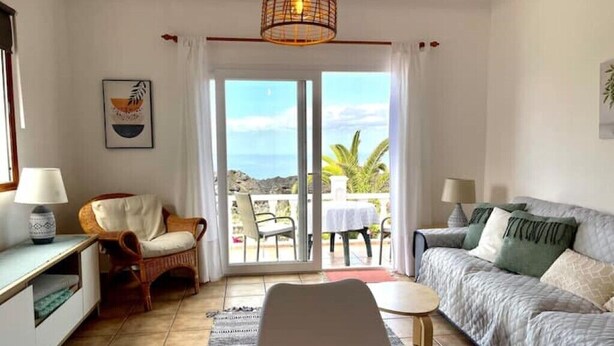 A room with a sofa, a chair, a table, and a view of the sea through a glass door.