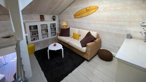 A loft-style room with a high ceiling, a white sofa with cushions, a small white stool, a black rug, and a wooden cabinet with shelves.