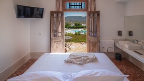 A bedroom with a bed, a TV mounted on the wall, and a view of a pool and mountains through an open door. - Finca Maltés San José (Nijar)