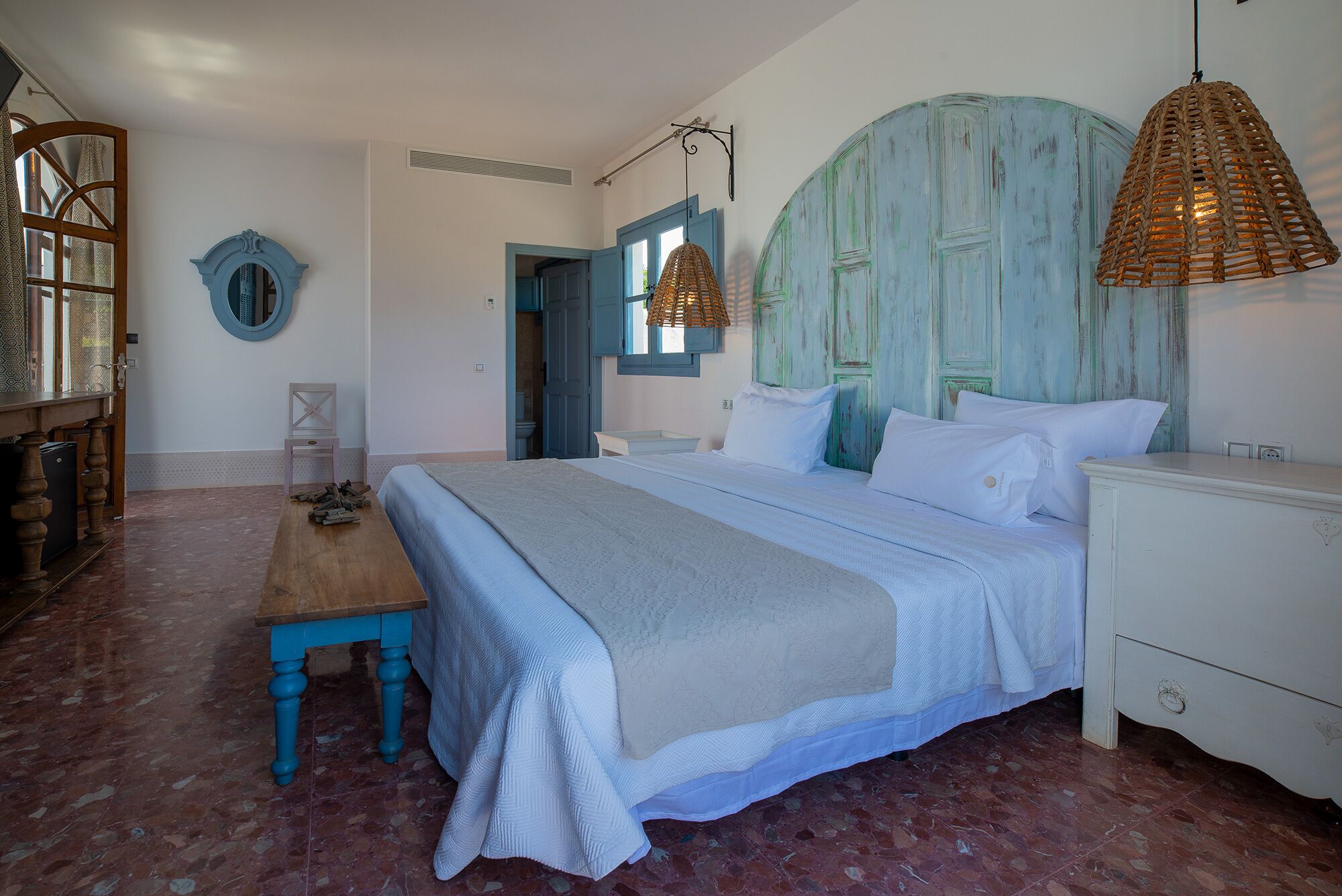 Un dormitorio con una cama grande, una mesita de noche de madera, una lámpara de mimbre y una puerta azul.
